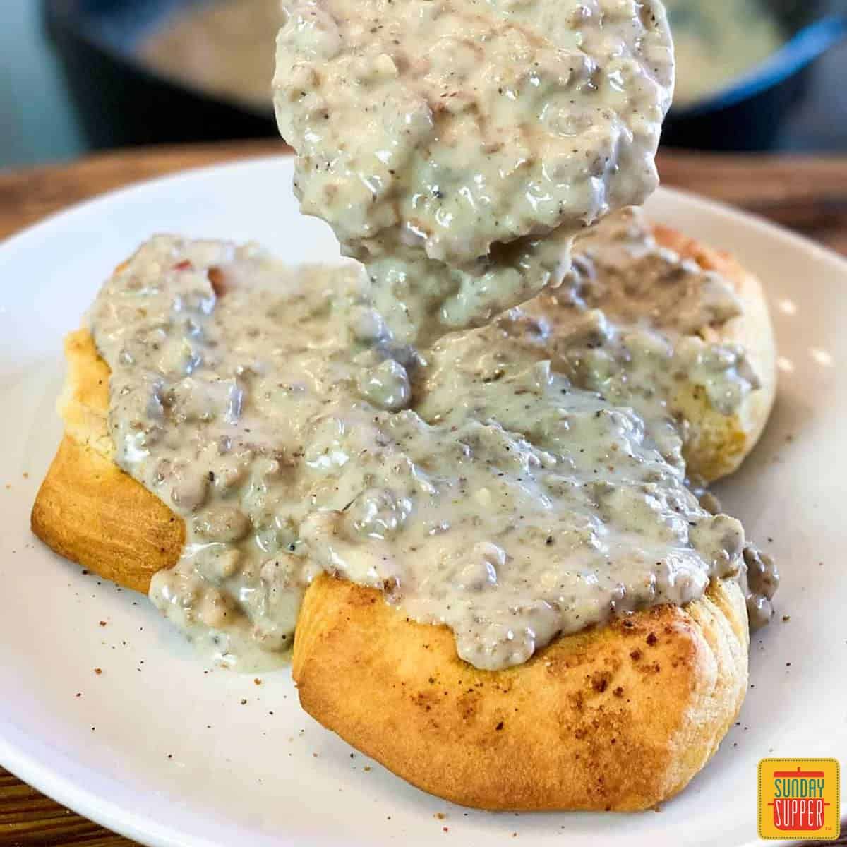 Spooning sausage gravy over biscuits on a white plate for breakfast for dinner