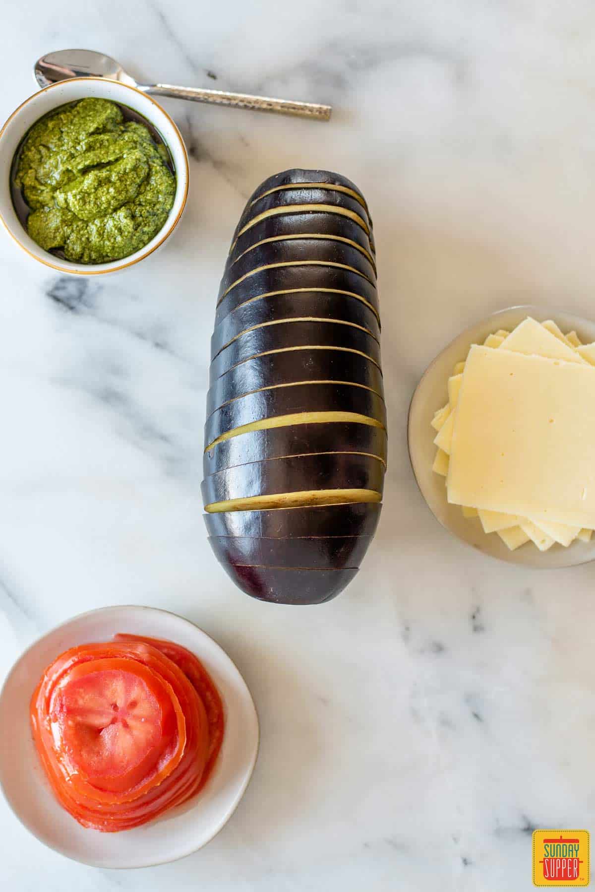 Pesto, tomatoes, and cheese in bowls next to an eggplant that's been sliced