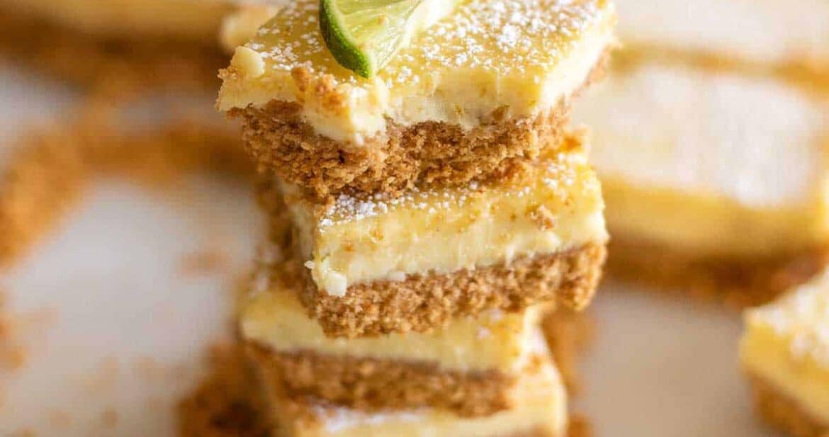 Three key lime pie bars stacked on top of each other surrounded by cookie crumbles with a lime slice on top