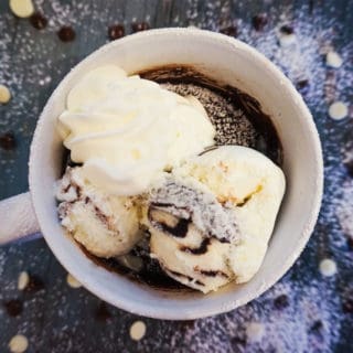 Brownie in a mug with whipped cream and ice cream on a table with chocolate chips spread around