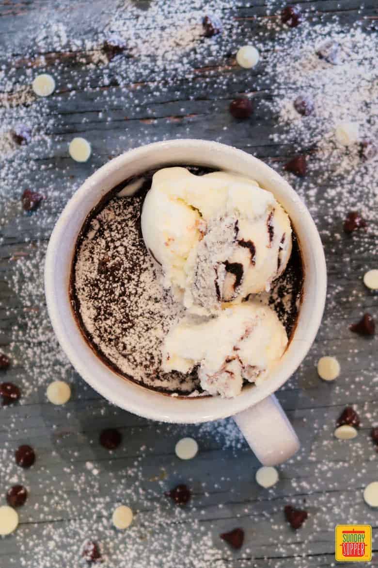 Brownie in a mug on a table surrounded by chocolate chips with whipped cream and ice cream topping