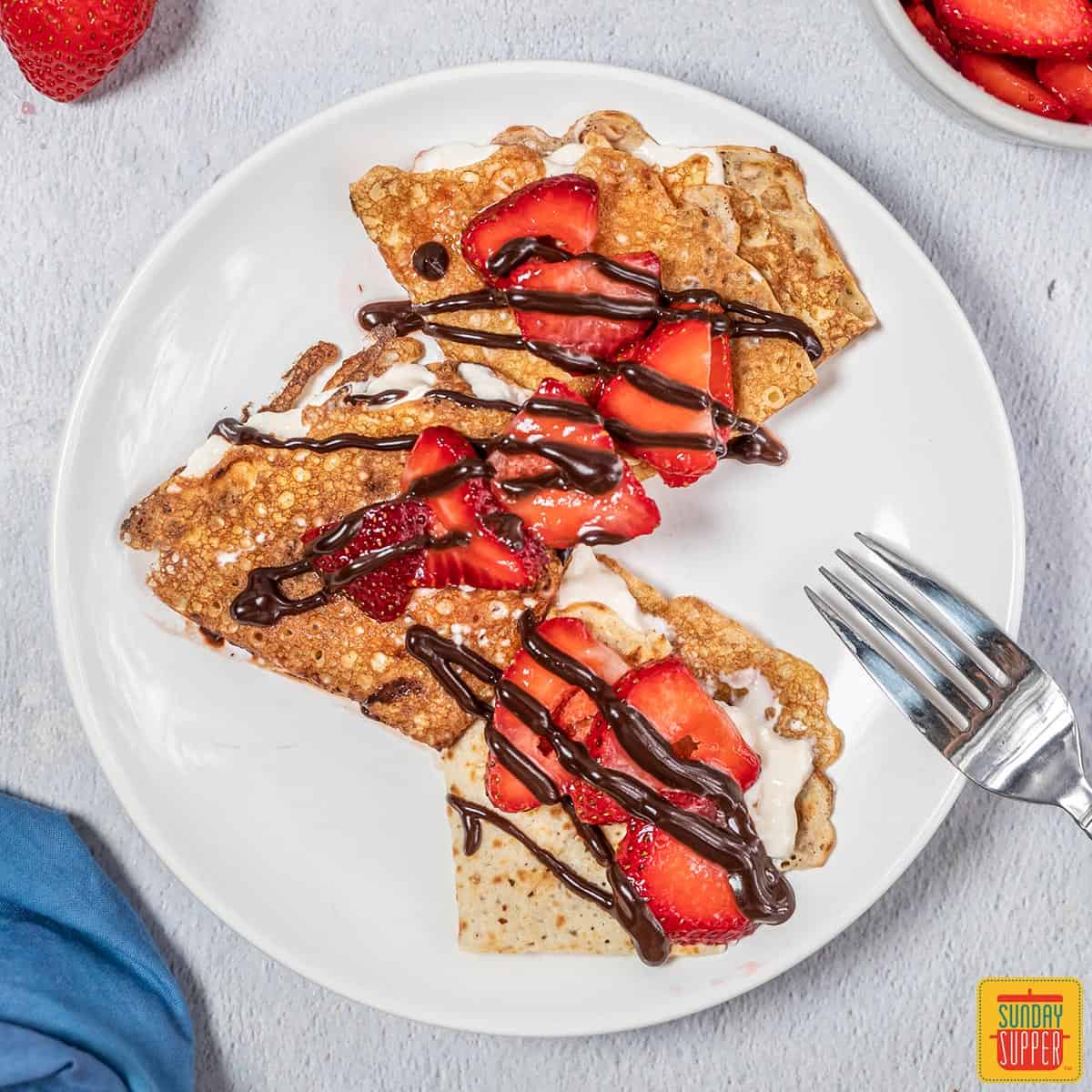 Three strawberry crepes on a plate with chocolate sauce
