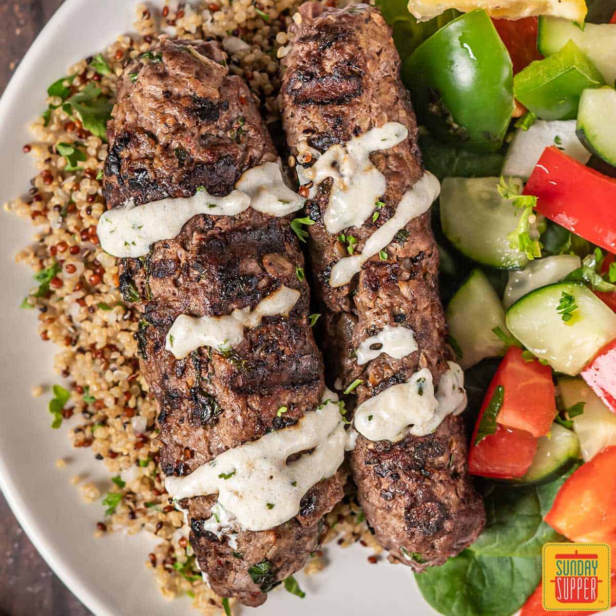 Beef kofta on a plate with cream sauce, couscous and a cucumber salad