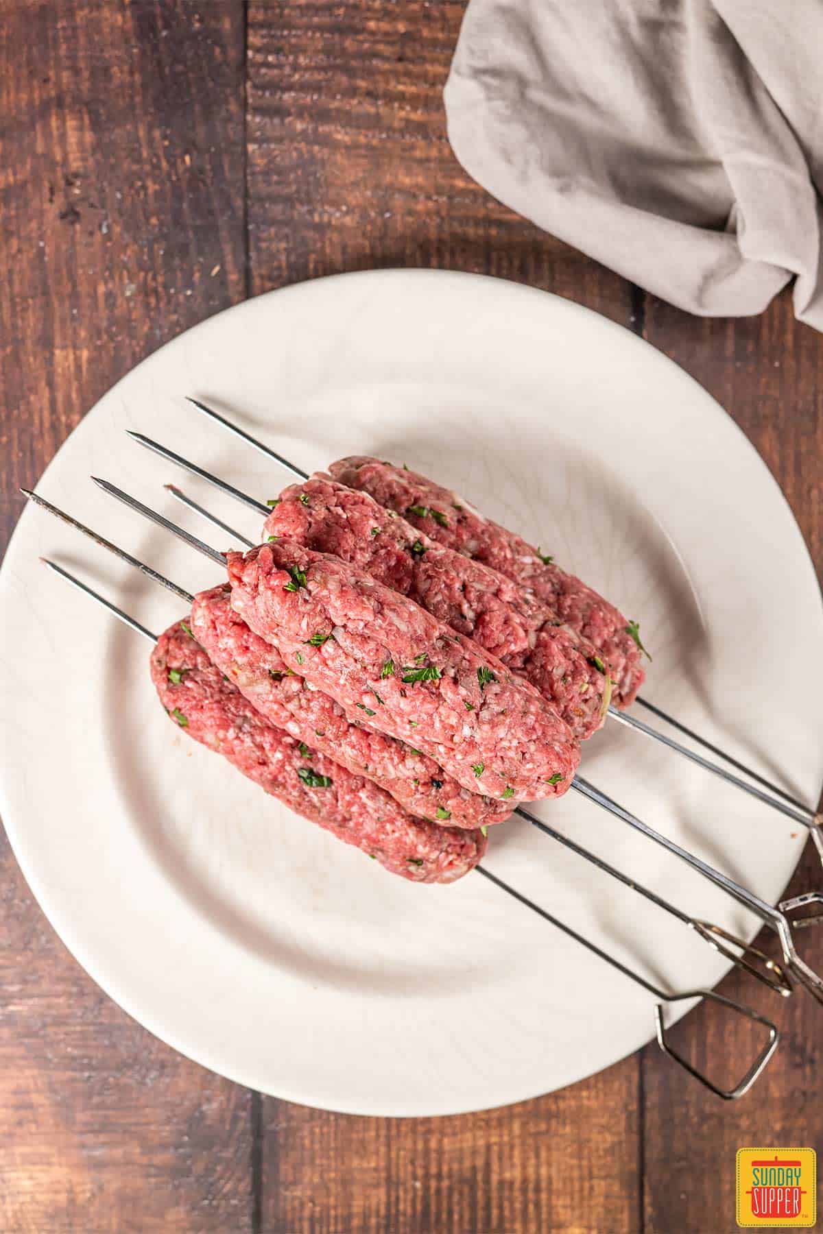 Uncooked lebanese kebabs on a white plate on metal skewers