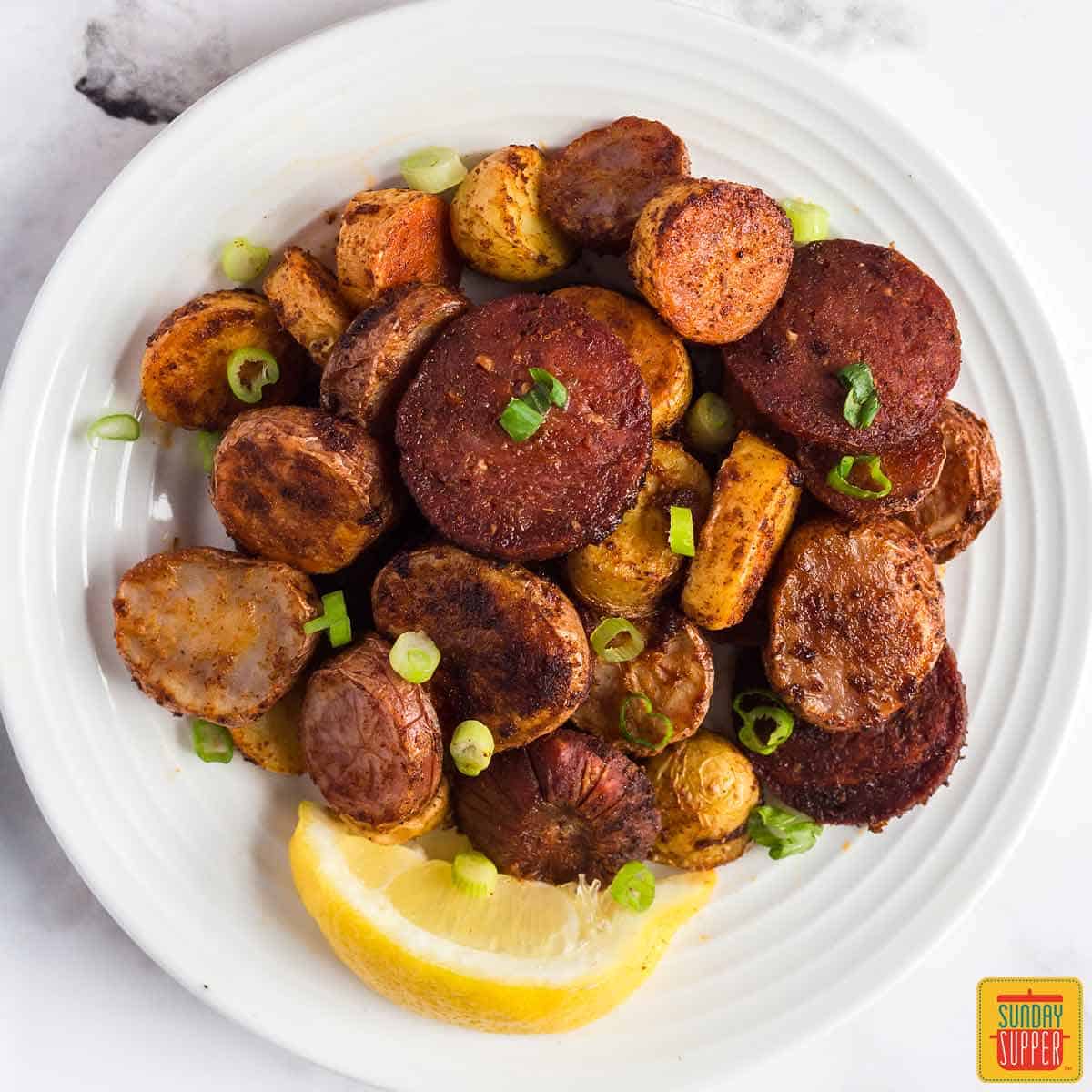 A white plate of Portuguese potatoes and chorizo with a lemon wedge