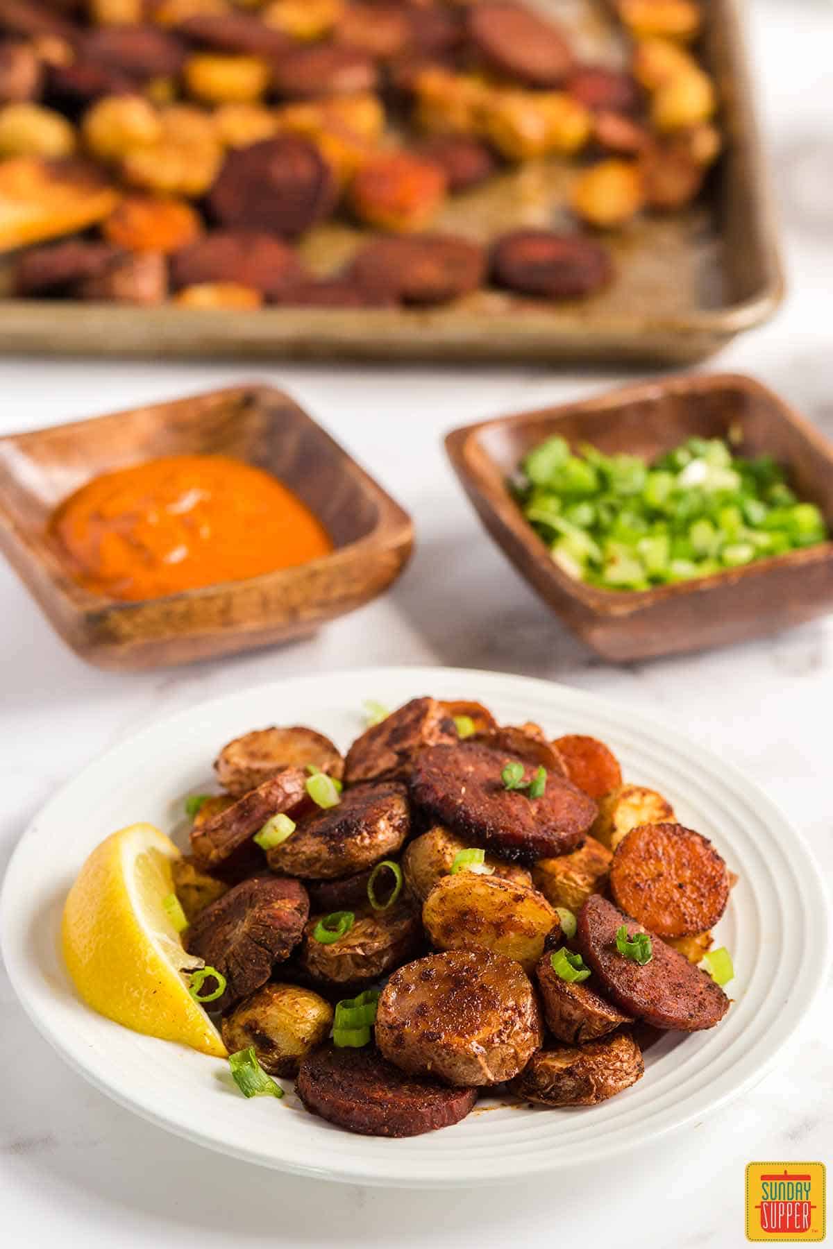 Portuguese potatoes and chorizo on a white plate with a lemon wedge next to bowls of peri peri sauce and green onions