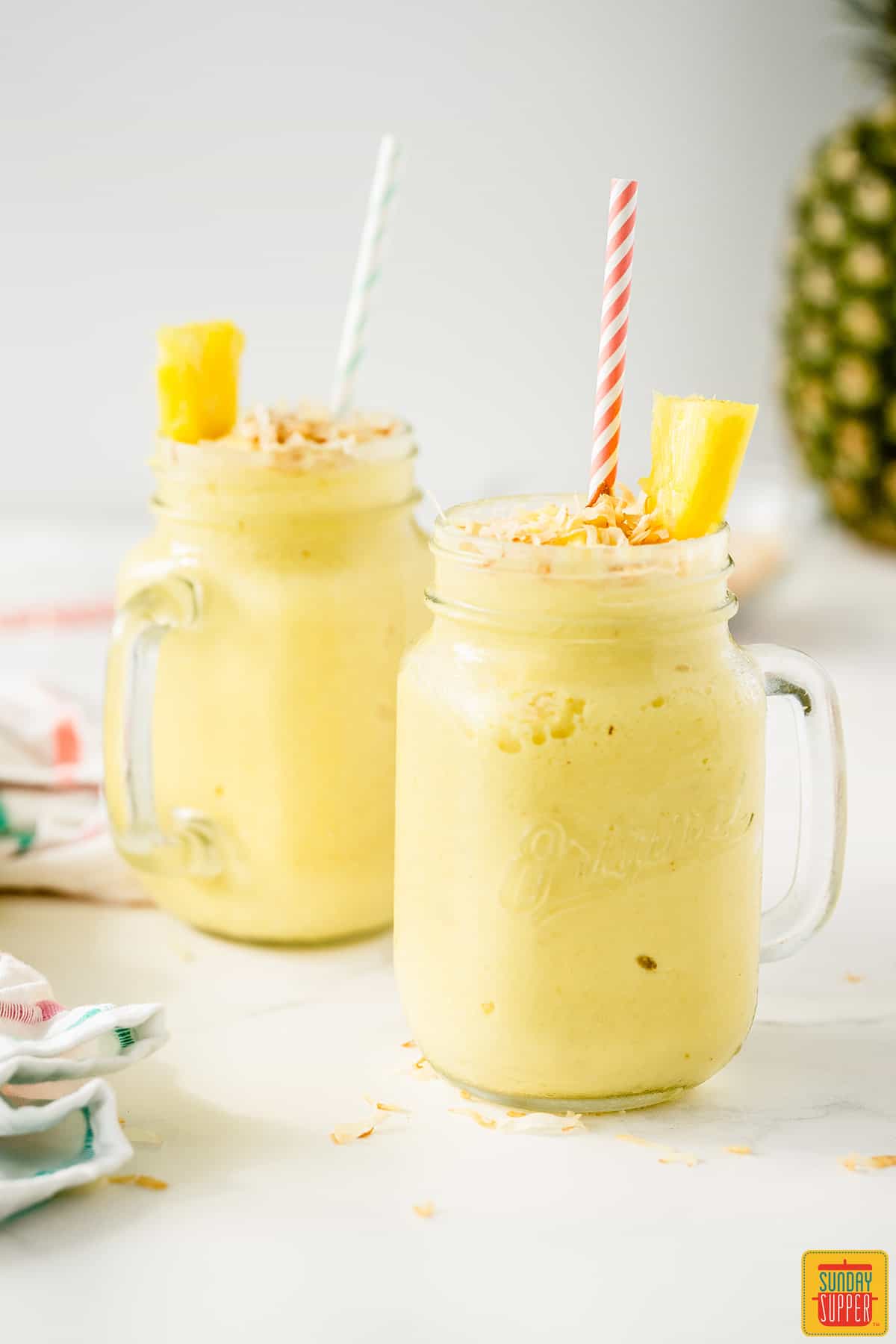 Coconut rum cocktail in two mason jar glasses with straws, pineapple slices, and toasted coconut flakes