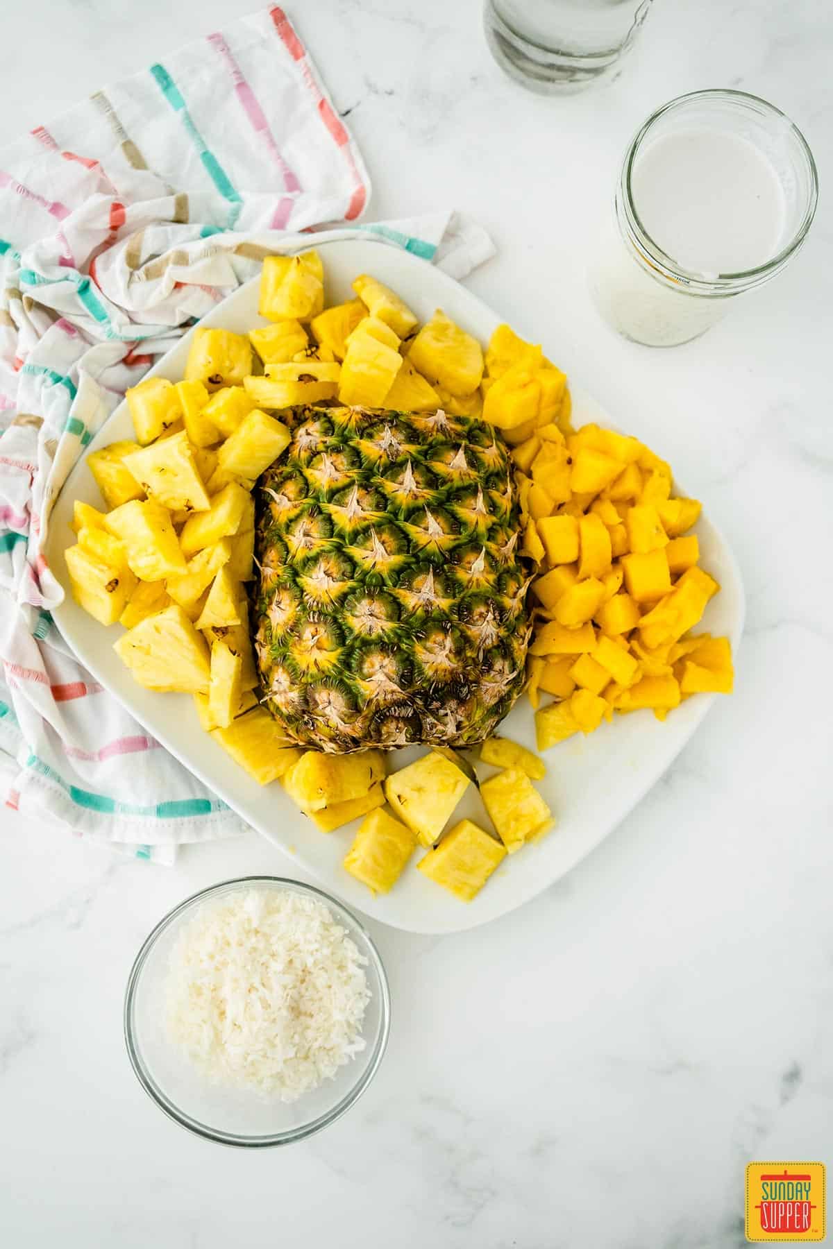 Half a pineapple surrounded by pineapple chunks and mango on a white platter with a bowl of coconut flakes and a glass of coconut milk to the side