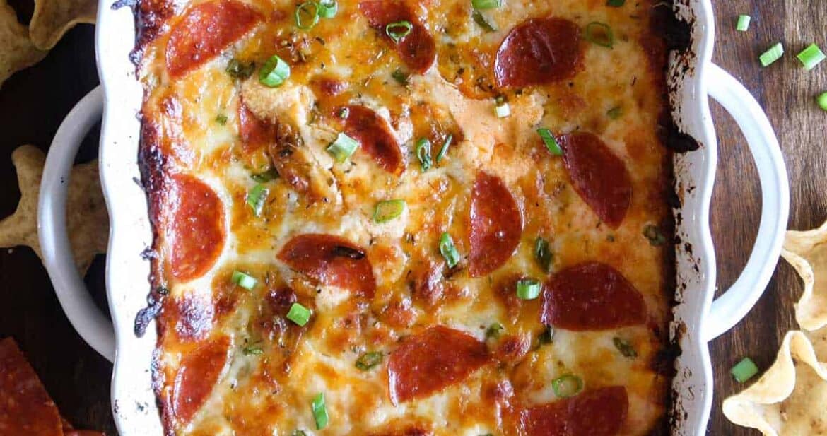 Pepperoni pizza dip in a white dish surrounded by pepperoni, chips, and onions