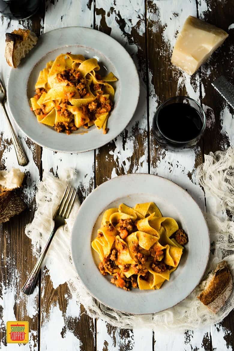 Two off-white plates of pappardelle pasta with homemade bolognese sauce on a distressed wooden surface with a fork, crusty bread, and red wine in a glass