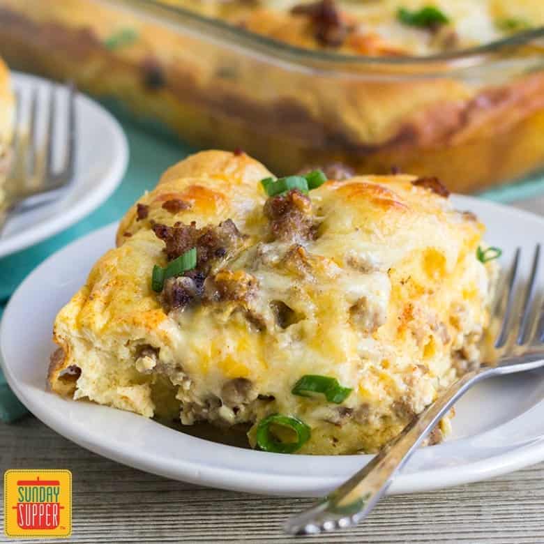 A slice of sausage breakfast casserole with crescent rolls on a plate with a fork in front of the casserole dish