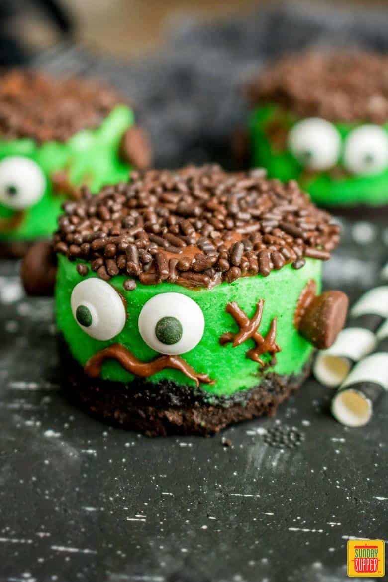 Mini halloween cheesecakes on a black surface decorated like Frankenstein with candy eyes