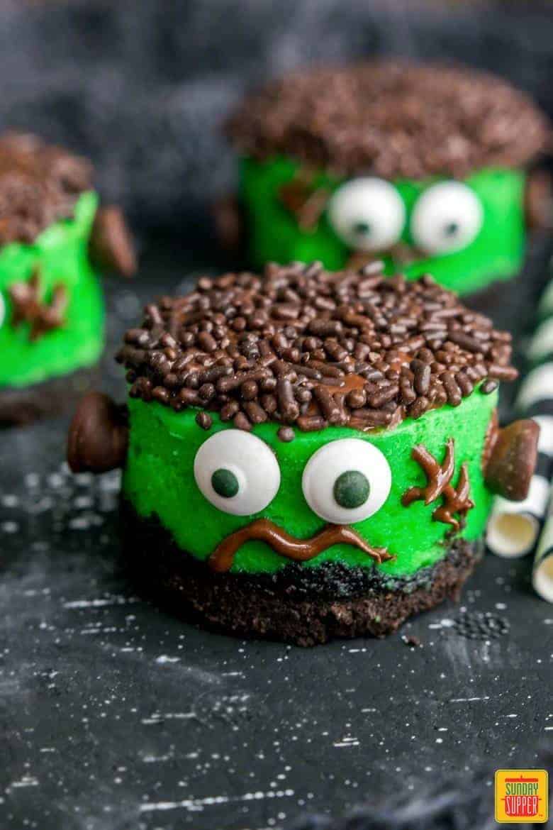 Mini cheesecakes for Halloween on a black surface - three decorated like Frankenstein with green frosting and candy eyes
