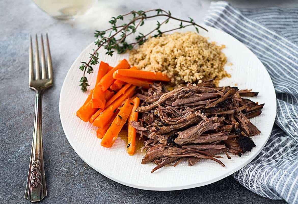 A white plate of slow cooker roast beef with glazed carrots and couscous