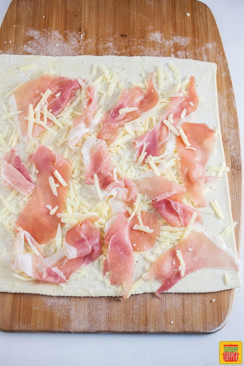 Sliced prosciutto and gruyere added to pinwheels on a wooden serving board