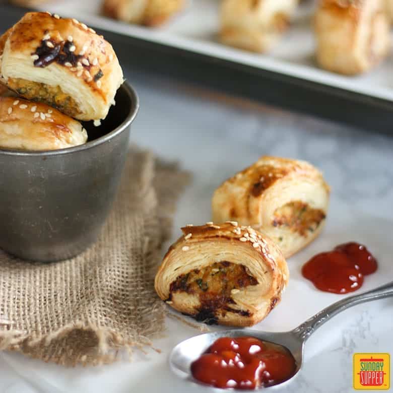 Four sausage rolls: two on the table next to a spoon with ketchup, and two in a metal tin next to them, with more visible in the background