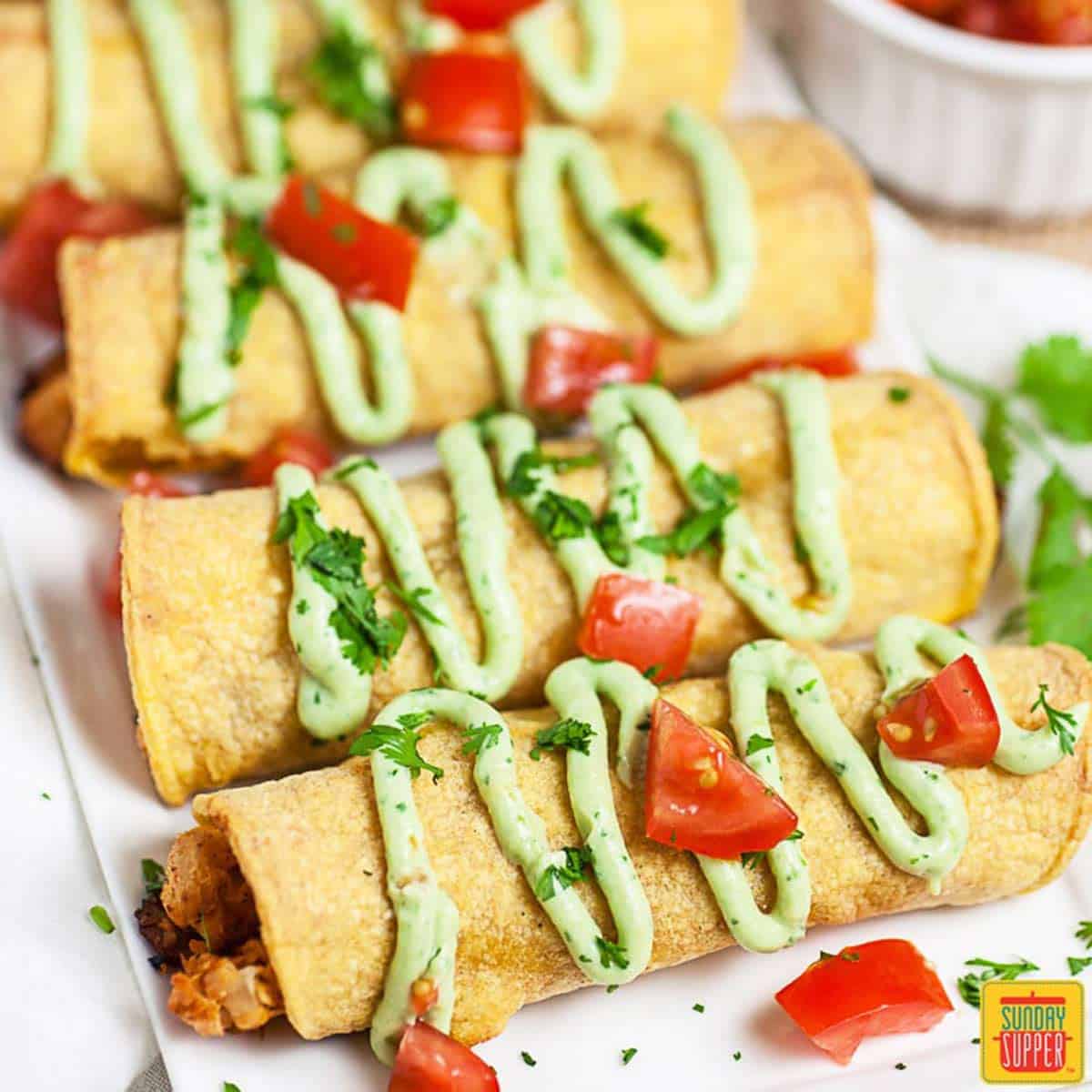 Row of 4 chicken taquitos with avocado crema and fresh tomatoes