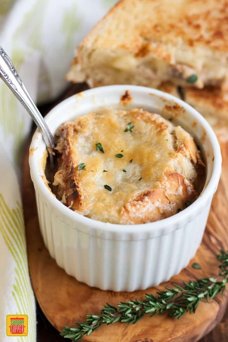 A ramekin of french onion soup with a spoon ready to eat and a mushroom grilled cheese sandwich on the side