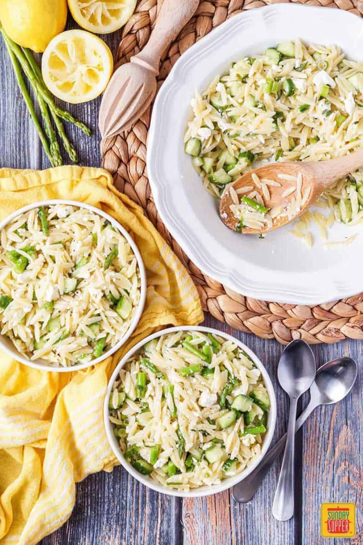 Two bowls of Lemon Orzo Pasta Salad next to a deep dish of pasta salad with a wooden serving spoon