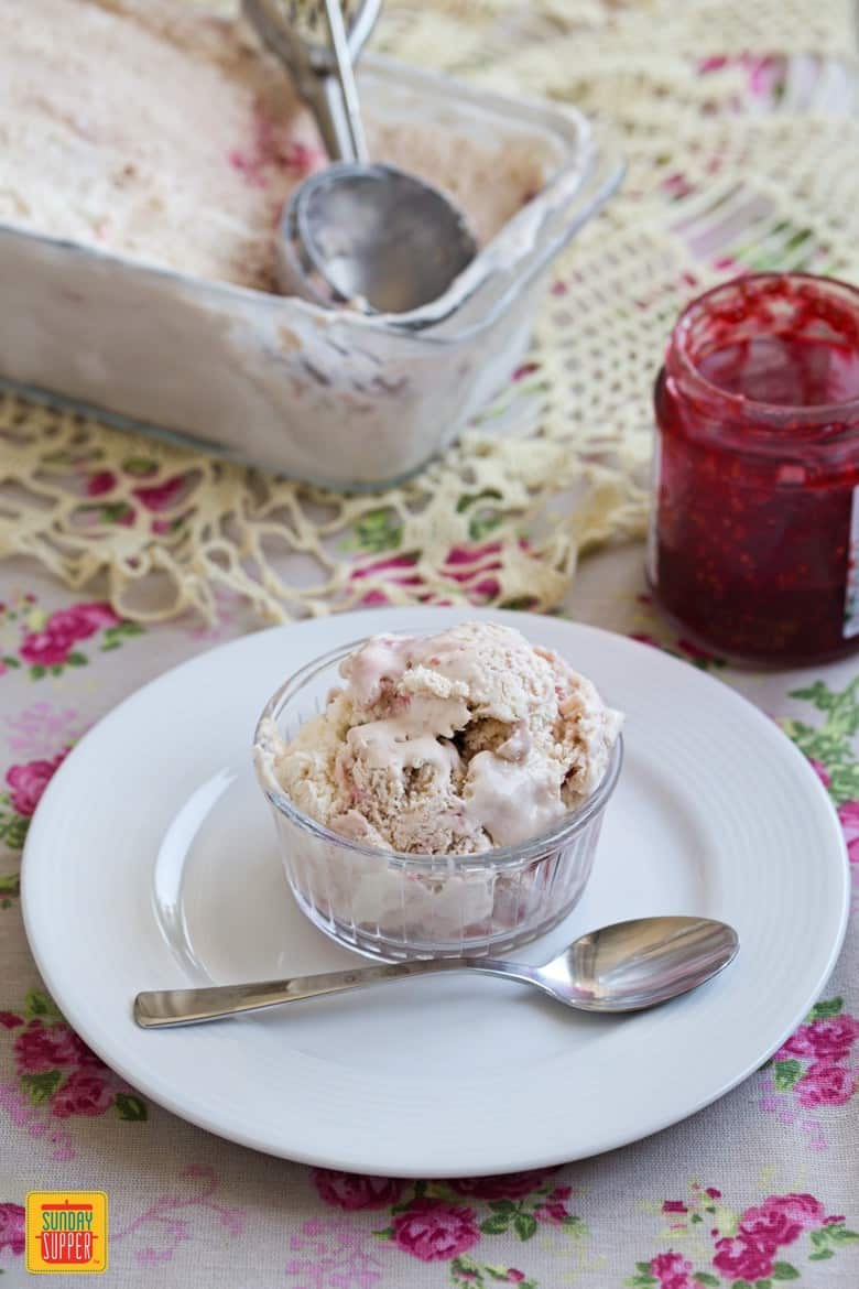Raspberry ice cream in a glass dish on a white plate with raspberry jam in a jar to the side and a spoon