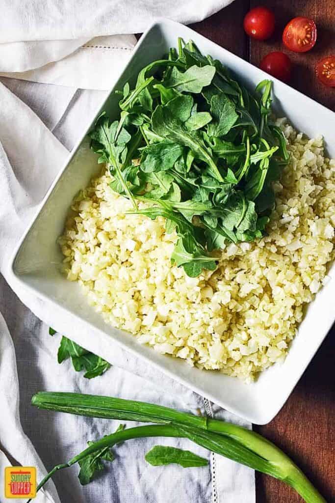Assembling the Cauliflower Rice Salad starting with baby arugula on top of the cauliflower rice