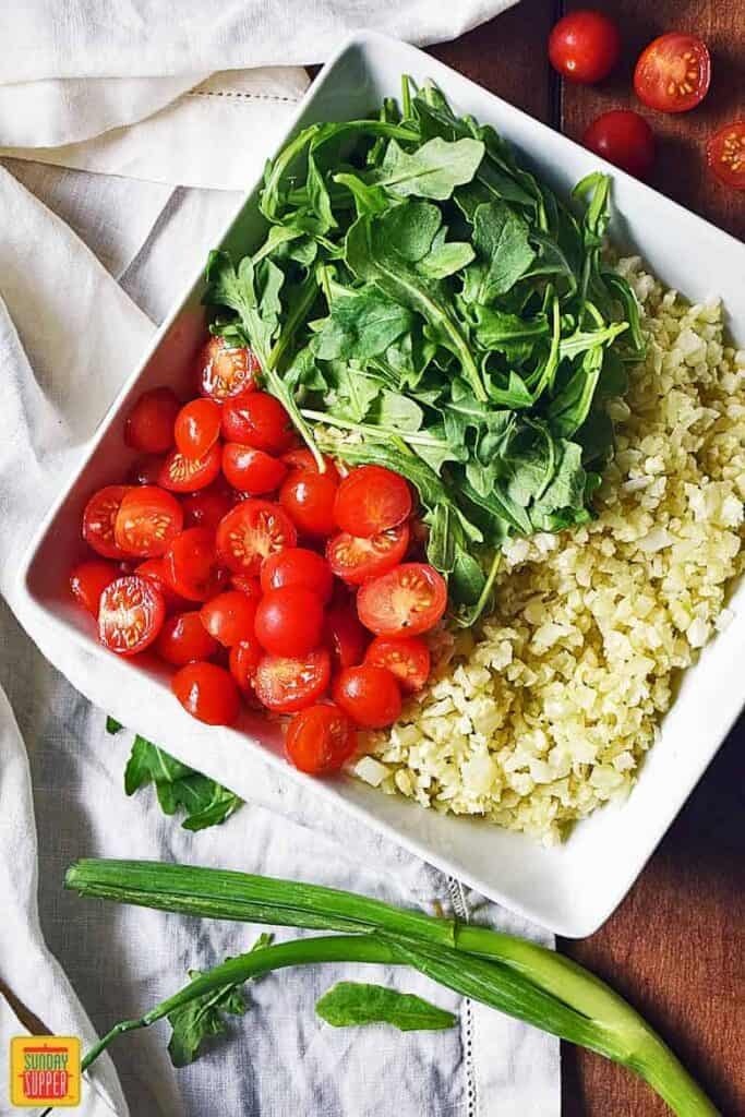Assembling the Cauliflower Rice Salad with baby arugula and halved tomatoes on top of the cauliflower rice
