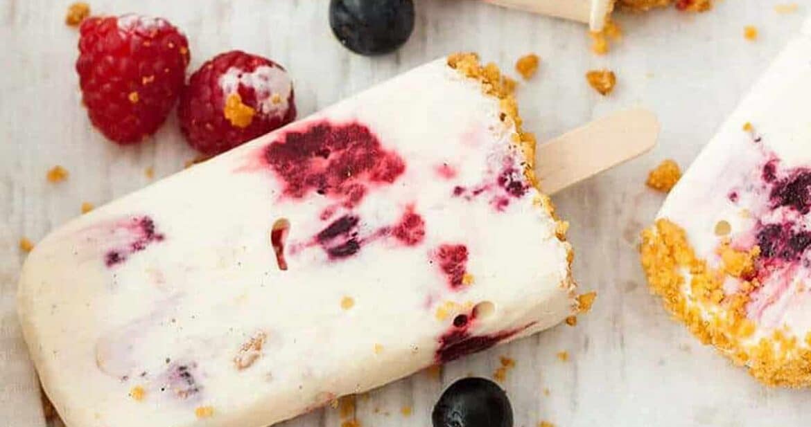Frozen cheesecake popsicles on a white surface with blueberries and raspberries