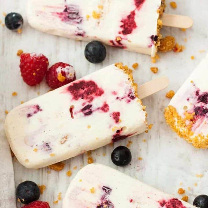 Frozen cheesecake popsicles on a white surface with blueberries and raspberries