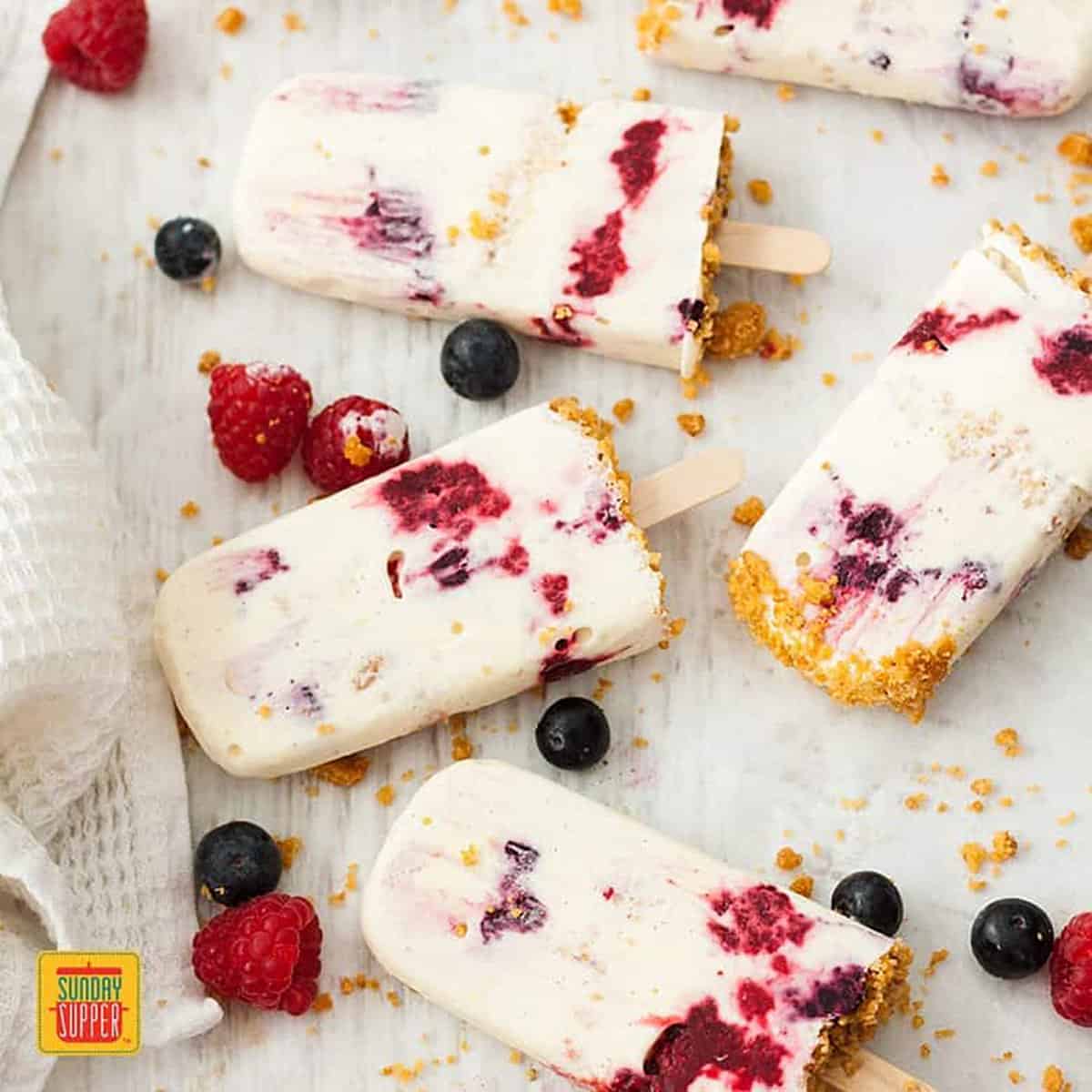 Frozen cheesecake popsicles with whole blueberries and raspberries on a white wood surface