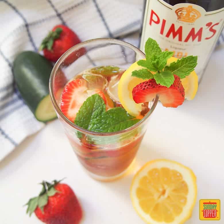 Pimm's Cup Cocktail in a tall glass next to a bottle of pimm's, with a lemon slice and strawberry on the rim of the glass