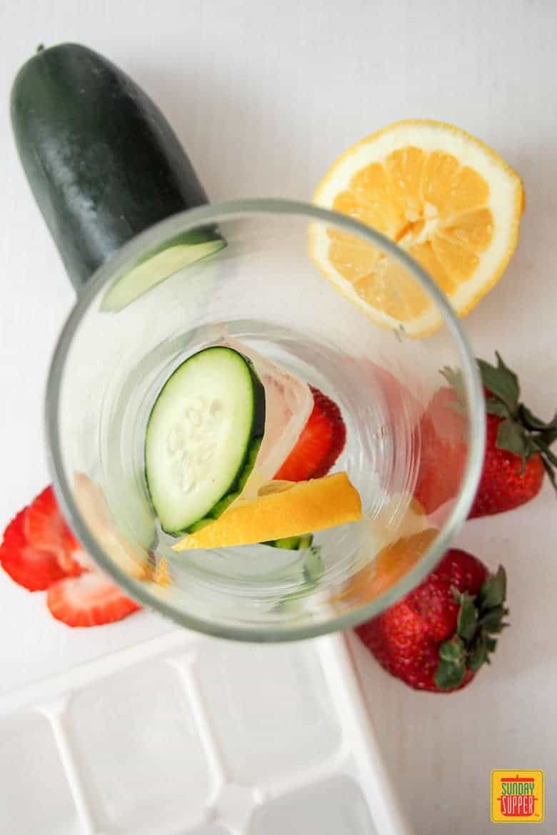Making the pimm's cup cocktail by adding a cucumber slice, ice, strawberry, and lemon to a glass