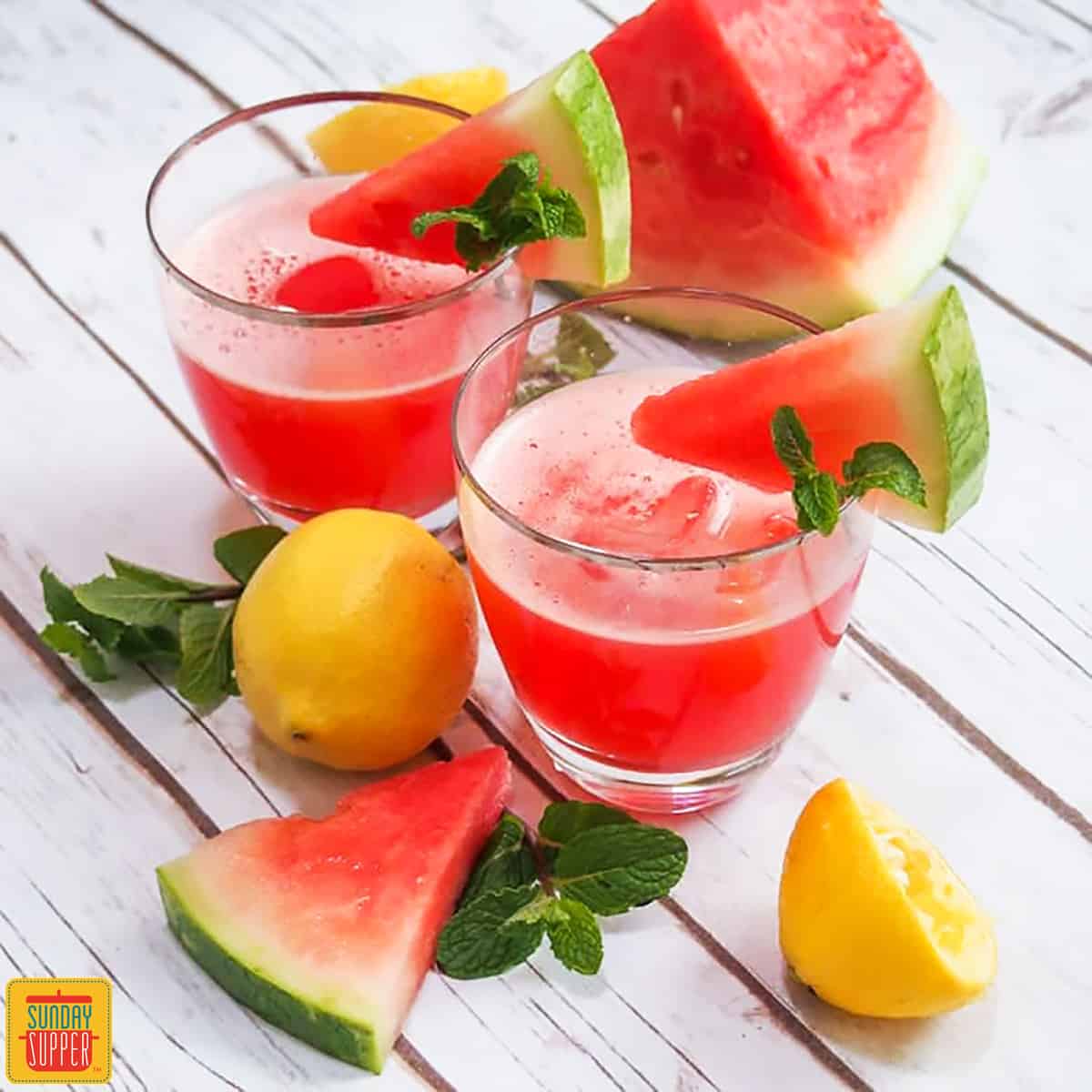 Two glasses of watermelon lemonade with watermelon slices and a chunk of watermelon in the background