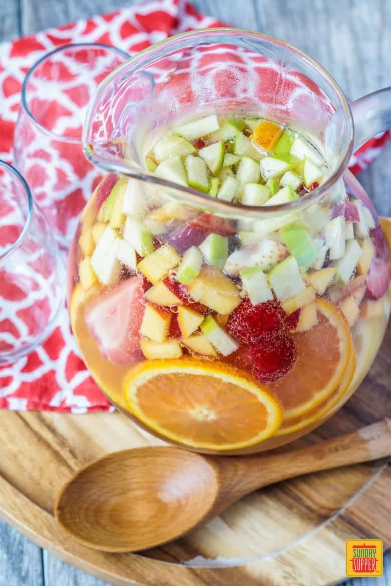 Moscato Sangria in a pitcher with fruit slices and a wooden spoon next to a couple of empty glasses