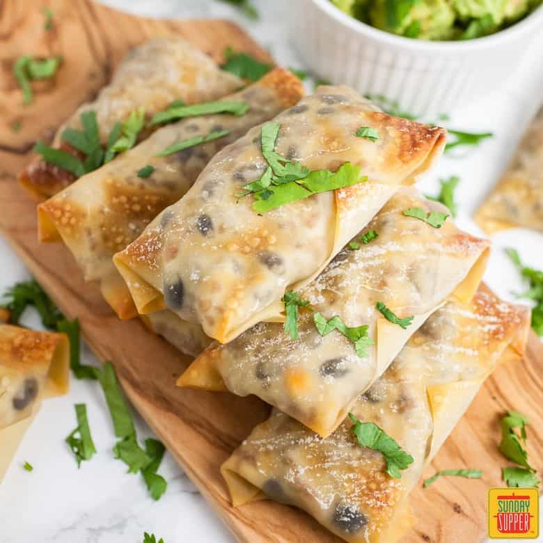 Baked Southwest Egg Rolls all stacked up on a wooden serving board with guacamole on the side in a ramekin