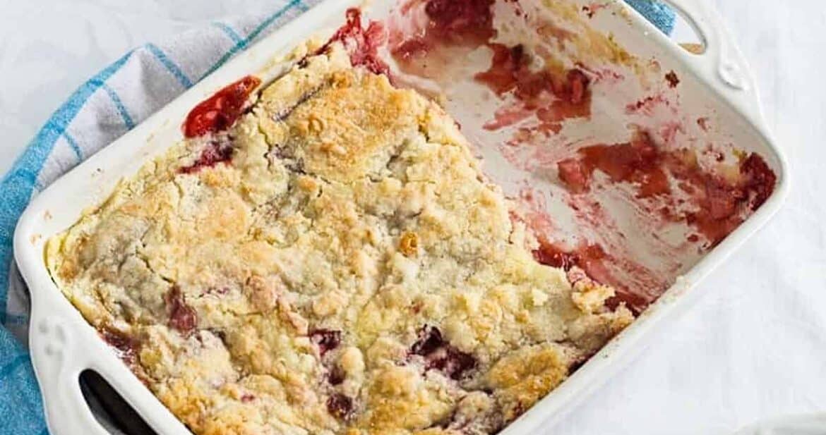 Strawberry dump cake with bananas in a white baking dish over a blue towel on the table with two plates of dump cake slices visible