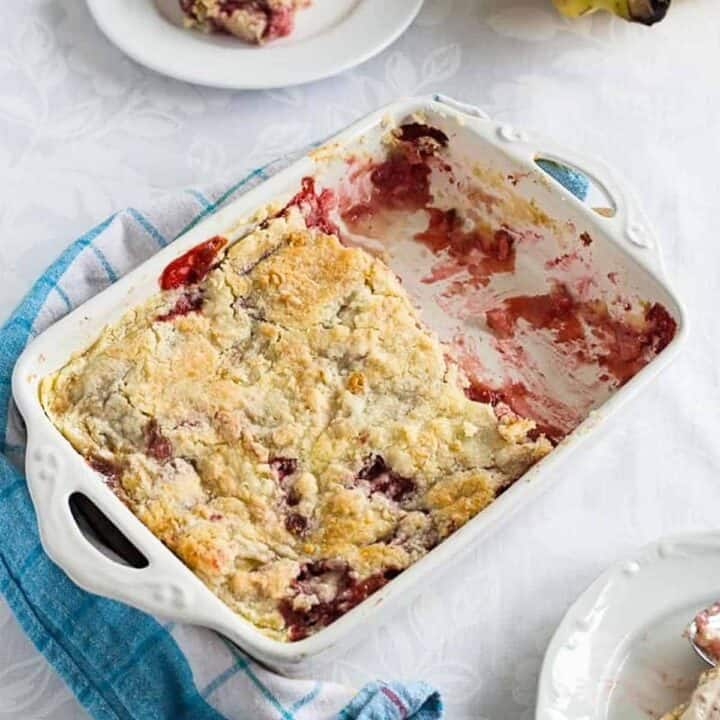 Strawberry dump cake with bananas in a white baking dish over a blue towel on the table with two plates of dump cake slices visible