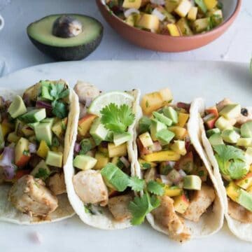 four fish tacos on a marble and wood board, half an avocado and bowl of salsa in background