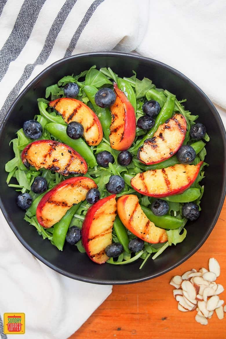 adding blueberries to grilled peaches salad in a black bowl