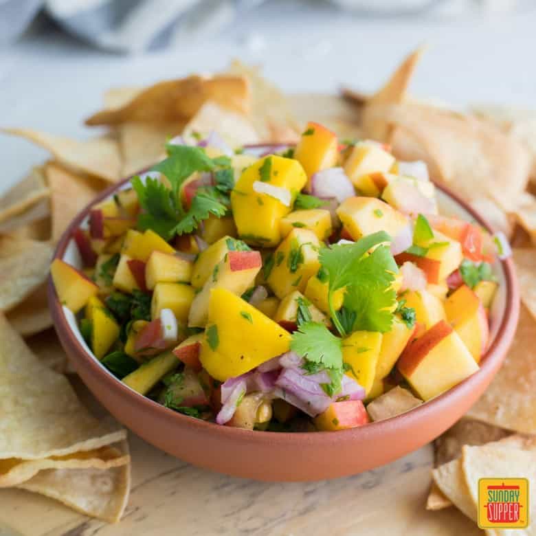 bowl of peach mango salsa on wooden round board surrounded by tortilla chips