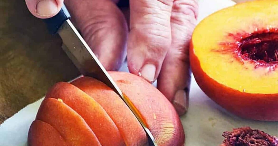 Cutting a peach into slices on a white surface next to half a peach and a peach pit