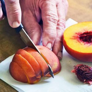 Cutting a peach into slices on a white surface next to half a peach and a peach pit