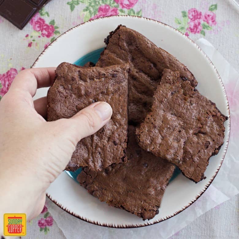 Gluten free fudge brownies stacked on a white plate with a hand reaching to pick one up