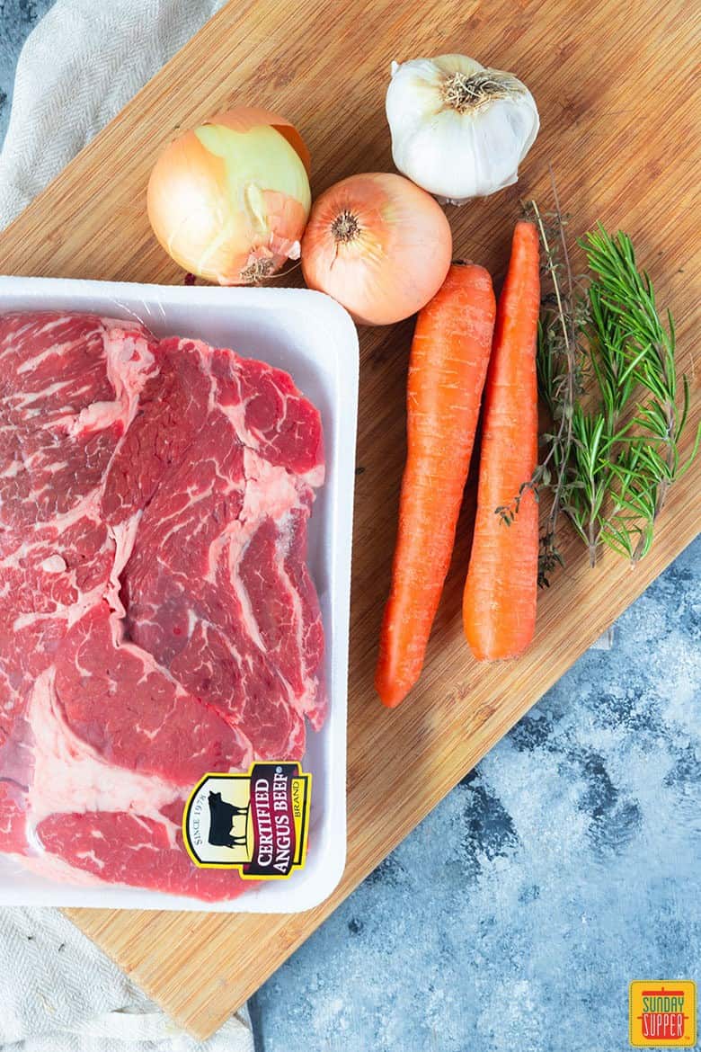 recipes to make dutch oven pot roast on a cutting board - chuck roast, onion, garlic, carrot and rosemary