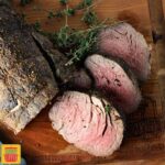 Whole beef tenderloin sliced on a cutting board with fresh sprigs of herbs