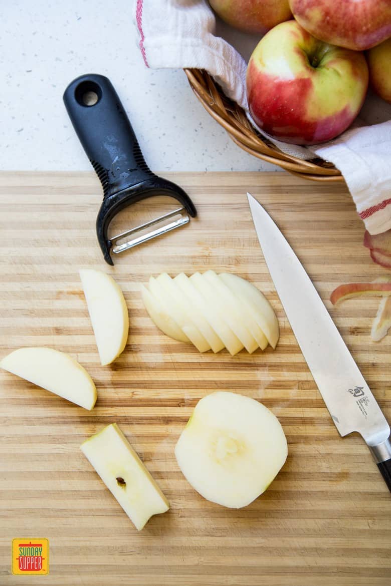 Slices of apples on a wooden cutting board next to a peeler and knife