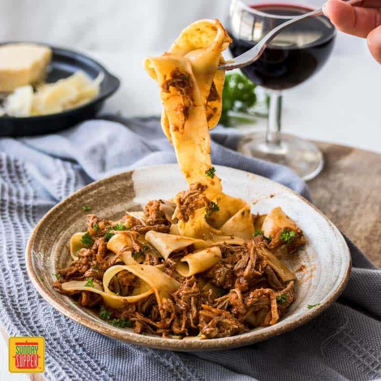 Slow Cooker Beef Ragu served over pappardelle pasta. A fork digging in with pasta dangling down to plate