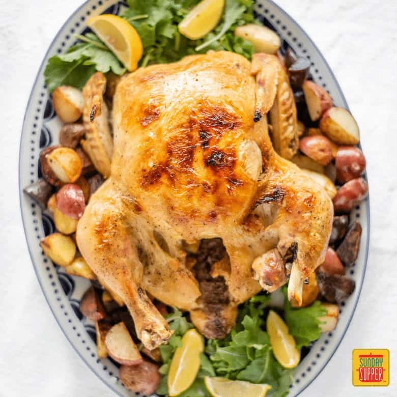 Crispy and juicy slow cooker whole chicken on a serving tray surrounded by greens, roasted potatoes, and lemon wedges