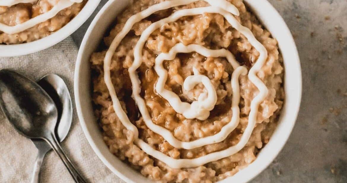 a bowl of slow cooker steel cut oats with cream cheese icing swirled on top. There's another bowl partially in view and two antique spoons sit atop a linen towel on the left hand side.