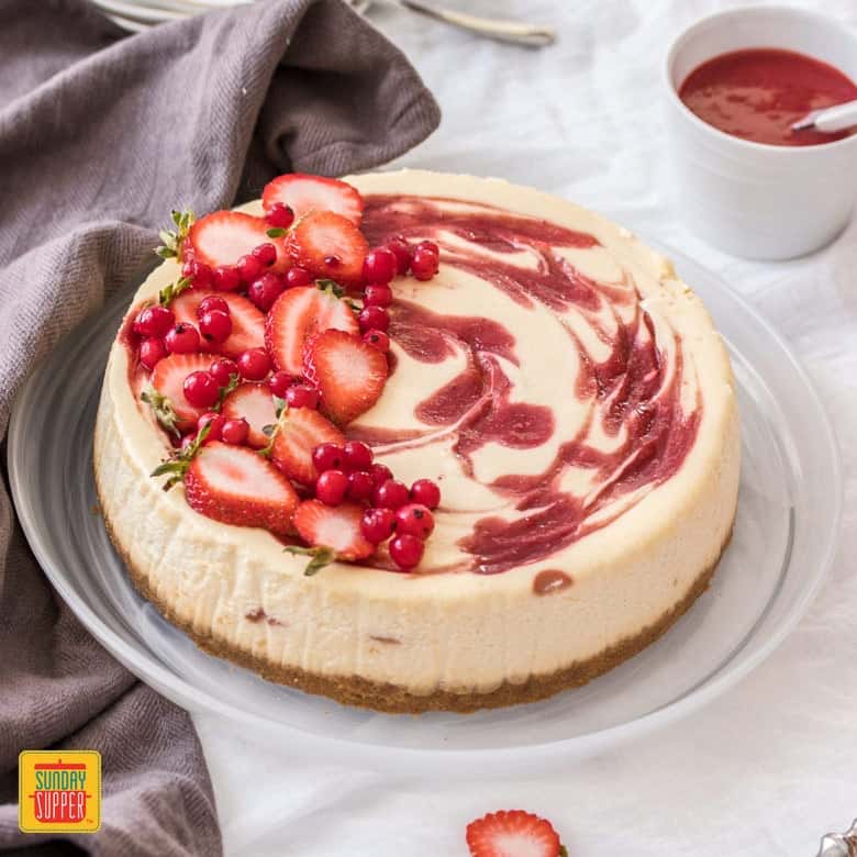 Whole strawberry swirl cheesecake on a glass platter withi strawberries on top