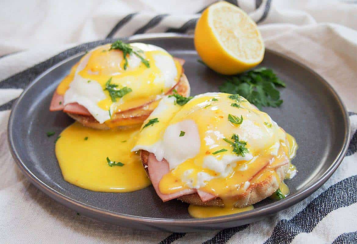 eggs benedict on a black plate with hollandaise sauce and half a lemon